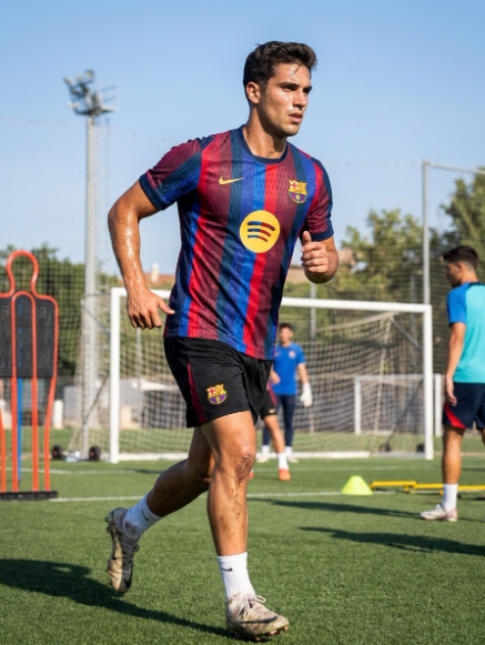 A soccer player in a red and blue striped FC Barcelona jersey with a yellow Spotify sponsor logo runs on a green turf field, with teammates and training equipment blurred in the background.